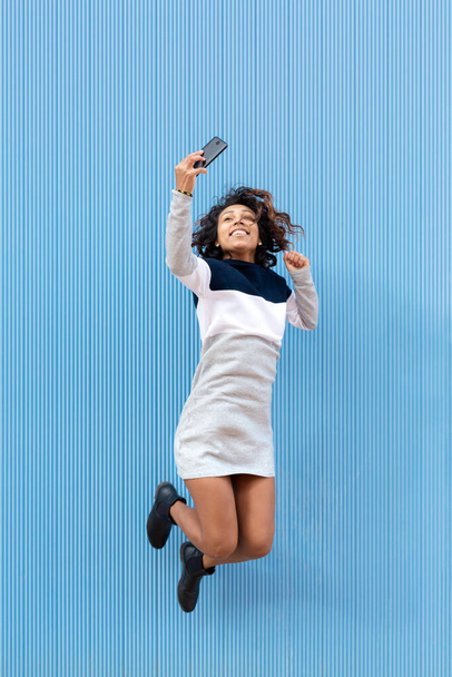Genç, gülümseyen bir kadın hoplayıp zıplıyor ve cep telefonu havada fotoğraf çekiyor. Mavi arka plan. Metin için boşluk. - Fotoğraf, Görsel
