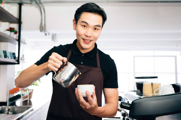 Junge männliche Barista blickt in die Kamera, trägt eine Schürze, die heiße Milch in heißen Espresso-schwarzen Kaffee gießt, um Latte Art zu machen. - Foto, Bild