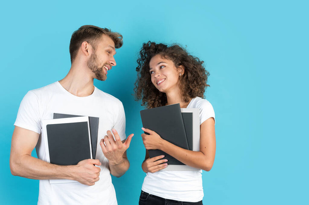 Study and education concept. Happy mixed race friends with exercise books talking to each other, smiling wide and standing on blue background with copy space, wearing in white t-shirts - Photo, image