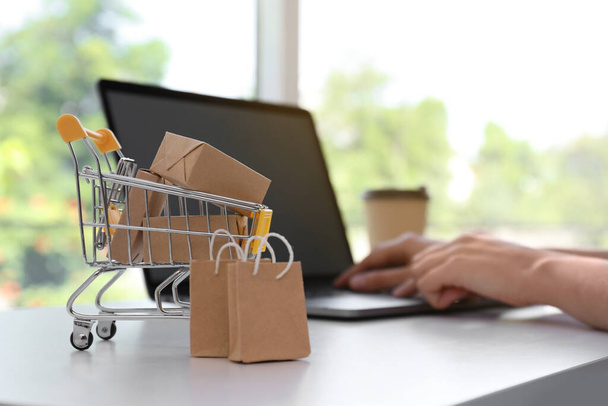 Internet shopping. Small cart with boxes and bags near woman using laptop indoors - 写真・画像