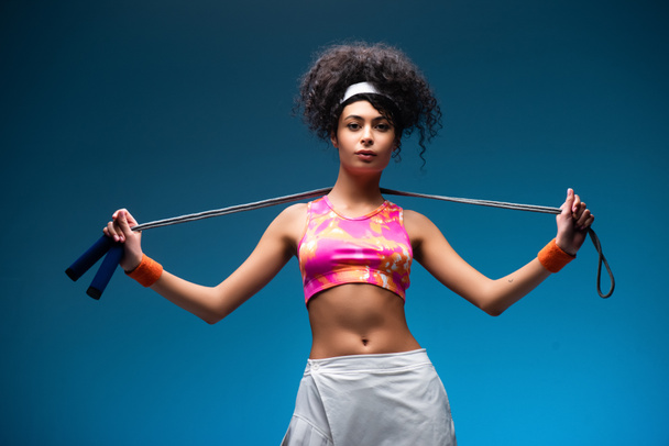 curly young sportswoman standing with skipping rope on blue - Foto, immagini