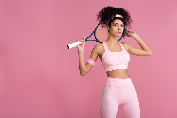 curly young sportswoman holding tennis racket while standing isolated on pink  - 写真・画像