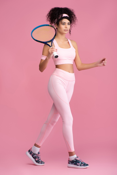 full length of young curly sportswoman holding tennis racket while walking on pink  - Foto, immagini