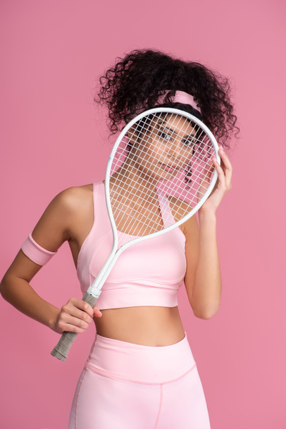 young sportive woman looking at camera through tennis racket isolated on pink  - Фото, изображение