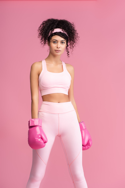 mujer joven y rizada en ropa deportiva y guantes de boxeo de pie aislado en rosa  - Foto, Imagen