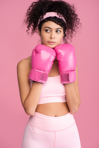σγουρά νεαρή γυναίκα σε αθλητικά ρούχα και γάντια πυγμαχίας κοιτάζοντας μακριά απομονωμένη σε ροζ  - Φωτογραφία, εικόνα