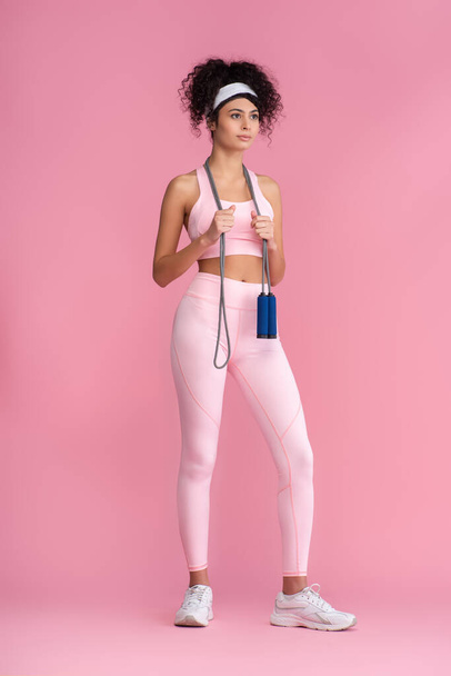 full length of curly young sportswoman standing with jumping rope on pink - Foto, imagen