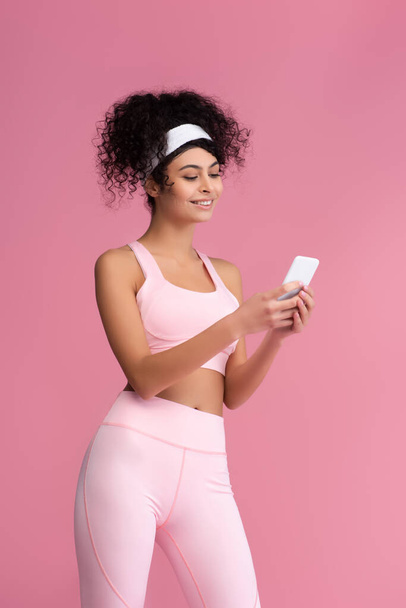cheerful young sportswoman using smartphone isolated on pink  - Foto, afbeelding