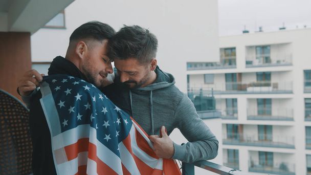 Happy handsome gay couple hugging under USA flag on the balcony - Photo, Image