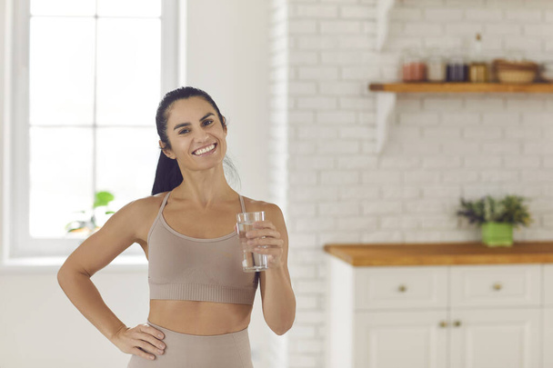 Lifestyle portrait of a fitness woman holding a glass of water while standing in the kitchen. - Foto, Imagen