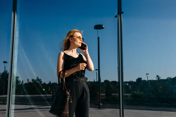 Şık giyinmiş güzel genç bir kadın şehir caddesinde telefonla konuşuyor. - Fotoğraf, Görsel