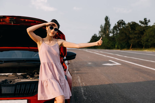 beautiful young woman with broken car showing hitchhikers gesture on road, having car trip during summer vacation - Foto, afbeelding