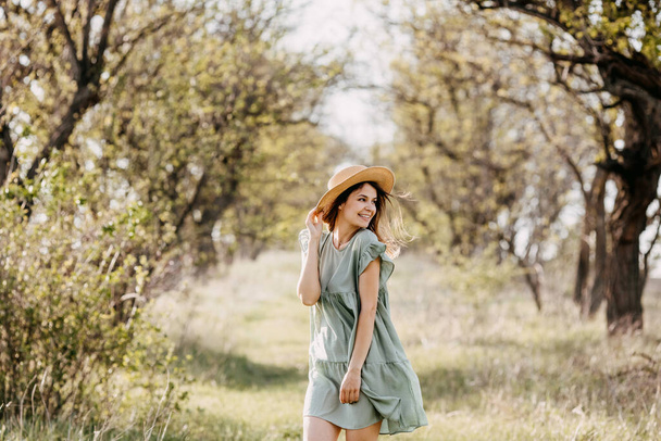 красивая молодая женщина в платье и соломенная шляпа в яблочном саду на закате - Фото, изображение