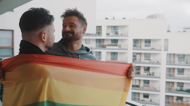 Νεαρό ενθουσιασμένο ζευγάρι γκέι που στέκεται στο μπαλκόνι καλυμμένο με σημαία ουράνιου τόξου - Πλάνα, βίντεο