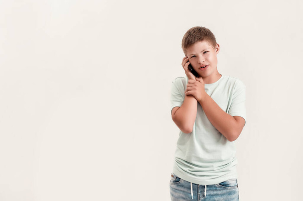 Portrait of teenaged disabled boy with Down syndrome looking at camera while talking on the phone, standing isolated over white background - Foto, imagen