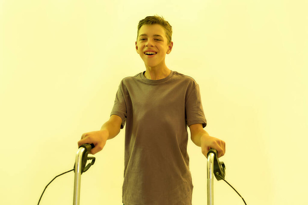 Cheerful teenaged disabled boy with cerebral palsy smiling at camera, taking steps with his walker isolated over yellow light background - Foto, Imagen
