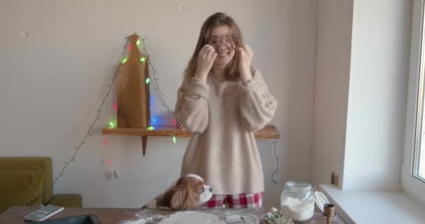 Vidám fiatal nő készül mézeskalács karácsonyra és újévre a konyhában a kutyájával, és a keksz vágó, mint a szemüveg. Lassú mozgás. - Felvétel, videó