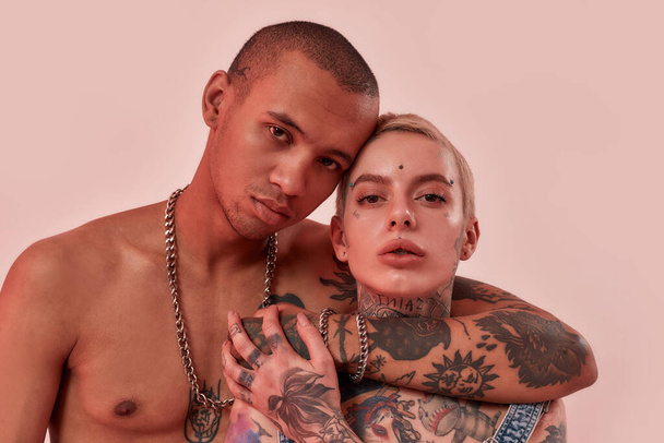 Dark-skinned topless male with tattoos hugging a pierced white tatooed woman, looking into a camera on a pinky background - Foto, imagen