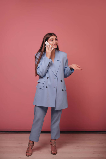 A smiling girl with long hair and in a blue business suit is talking on the phone on a pink background. - Photo, Image