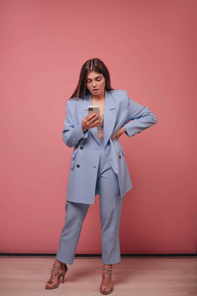 A girl with long hair and in a blue business suit looks at the smartphone screen on a pink background. - Fotó, kép