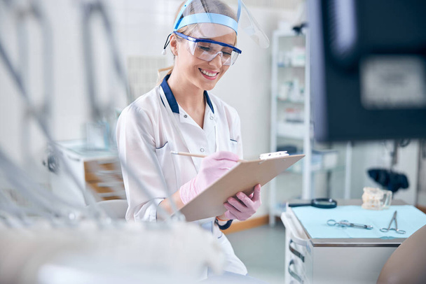 Dentiste féminine joyeuse en uniforme prenant des notes - Photo, image