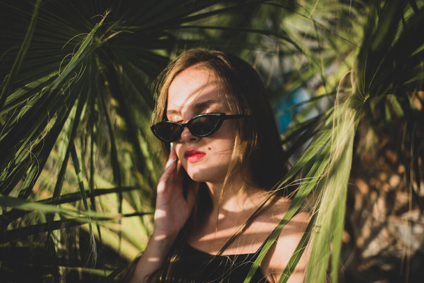 woman wearing sunglasses posing with palm tree. Portrait of beautiful teenage girl with shadows of palm leaf on her face - Фото, изображение