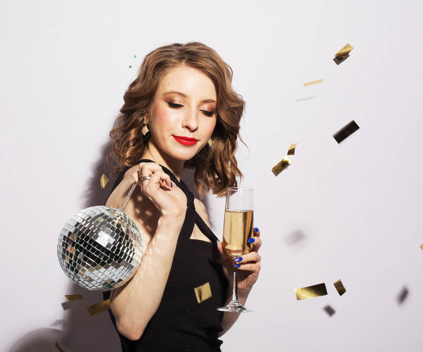 Beautiful women holding a glass of champagne and a disko ball on the background of falling sparkling confetti, party and holiday atmosphere - Foto, immagini