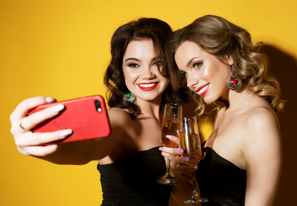Celebration, party, and people concept - two chic young women in black cocktail dresses drinking champagne and taking a selfie with mobile over yellow background - Photo, Image