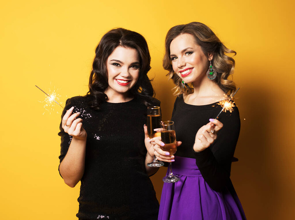 Holiday and party concept: Portrait of two young fashionable women with a champagne glasses and sparklers over yellow background - Foto, immagini