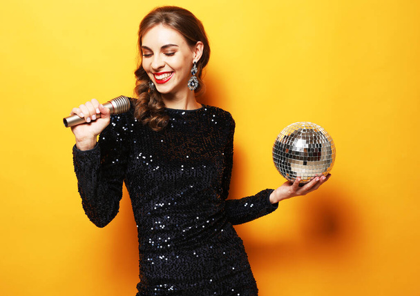 Party, holiday and celebration concept: Young brunette woman with long curly hair dressed in evening dress holding a microphone and disco ball, singing and smiling over yellow background - Zdjęcie, obraz