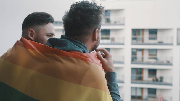 Молодая гомосексуальная пара наслаждается видом с балкона, покрытого радужным флагом - Фото, изображение
