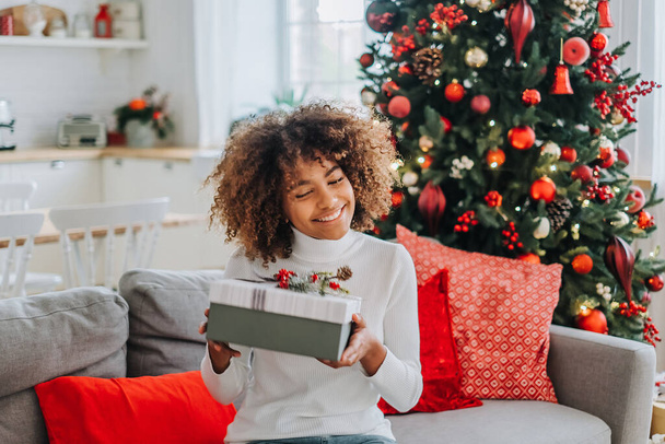 Piękna młoda Afroamerykanka otwiera prezent świąteczny w domu na kanapie na tle świątecznej choinki. - Zdjęcie, obraz