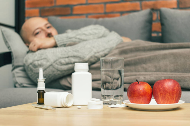 Concept of treatment virus infection in home, coronavirus, covid-19 or flu, table with medication, pills, water, apple in room and defocused sick man on sofa, guy is diseased and feels badly ill. - Fotó, kép