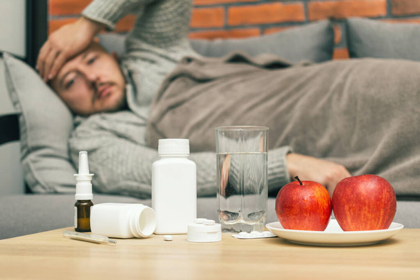 Concept of treatment virus infection in home, coronavirus, covid-19 or flu, table with medication, pills, water, apple in room and defocused sick man on sofa, guy is diseased and feels badly ill. - Foto, Imagen