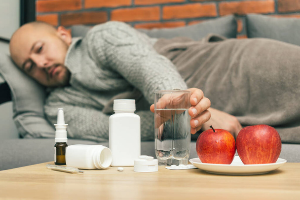 Concept of treatment virus infection in home, coronavirus, covid-19 or flu, table with medication, pills, apples in the room and defocused sick man on sofa takes glass with water. - Photo, Image