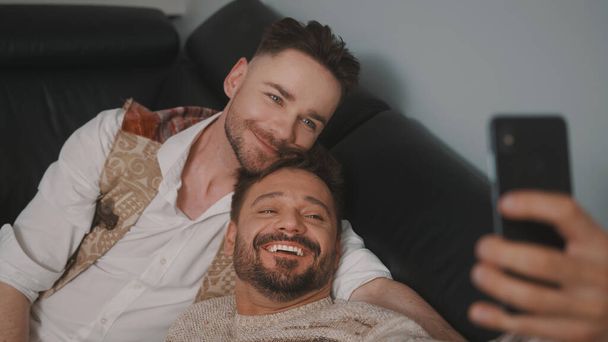 Ανδρικό ρομαντικό γκέι ζευγάρι χαλαρώνει στον καναπέ. Ο φίλος του φιλάει τον συνεργάτη του στο κεφάλι. Λήψη selfie με smartphone - Φωτογραφία, εικόνα