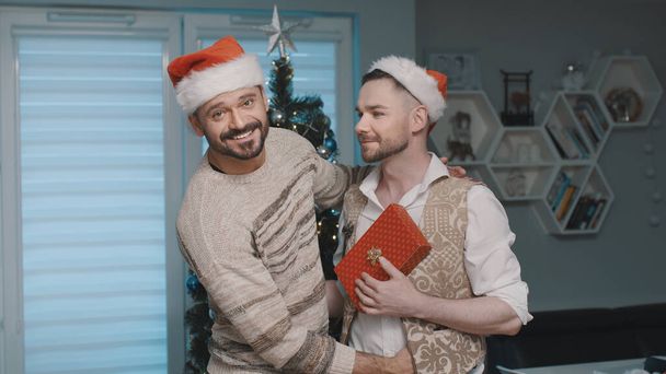 Kerstmis in gay familie, knuffelen in de voorkant van de kerstboom met geschenken en kerstman hoeden - Foto, afbeelding