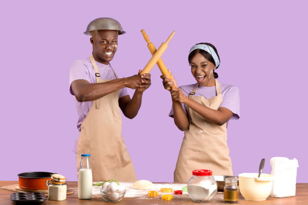 dwaas Afro-Amerikaans paar met mock vechten met rollende pinnen tijdens het bakken in de keuken, lila studio achtergrond - Foto, afbeelding
