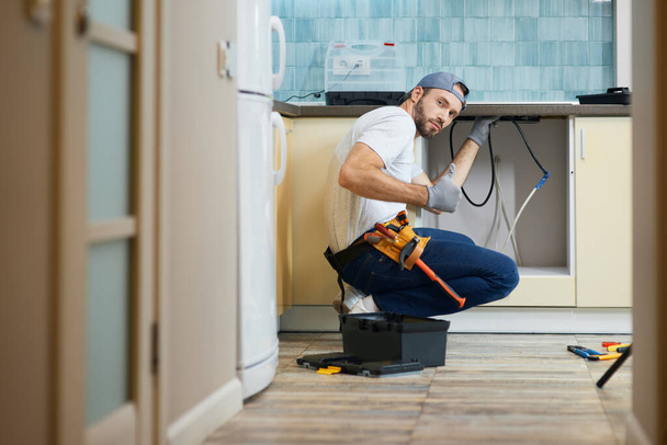 The work is done. Young repairman, professional plumber wearing tool belt looking at camera and showing thumbs up while crouching on the floor, fixing a pipe in the kitchen indoors - Foto, Imagem