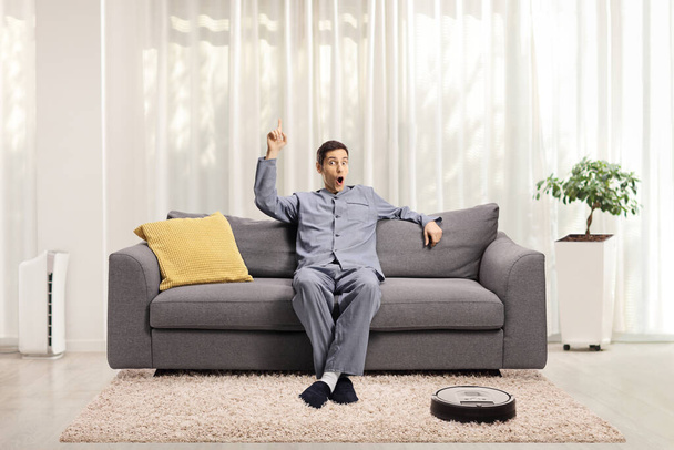 Νεαρός ενθουσιασμένος άνδρας με πιτζάμες κάθεται σε έναν καναπέ στο σπίτι δείχνοντας προς τα πάνω και μια ρομποτική ηλεκτρική σκούπα στο πάτωμα - Φωτογραφία, εικόνα