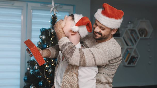 Hombres guapos felices con sombreros de Santa Claus abrazándose frente al árbol de Navidad. Regalo y relación homosexual - Foto, Imagen