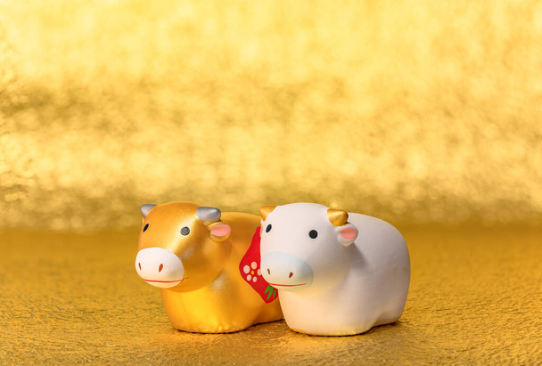 Grußkarte mit einer Nahaufnahme auf zwei niedlichen handgefertigten Figuren des Tierkreiszeichens von zwei Kühen, handbemalt in Gold und Weiß für das Jahr des Ochsen auf einem ungleichmäßig strukturierten goldenen Hintergrund. - Foto, Bild