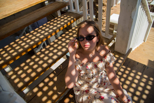 Um retrato de moda de uma bela jovem menina vestindo óculos de sol relaxando em uma escada em um dia ensolarado de verão no café na praia. jovem magro bela mulher férias de verão, ensolarado. - Foto, Imagem