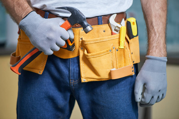 Listo para arreglar cualquier cosa. Primer plano del reparador con un cinturón de herramientas con varias herramientas, sosteniendo un martillo en su mano - Foto, Imagen