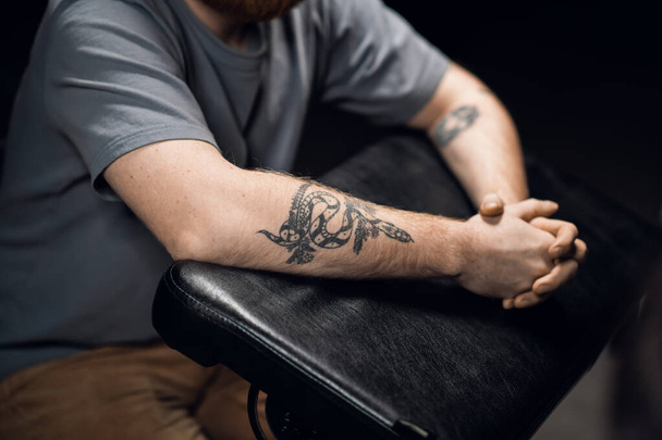 Κοντινό πλάνο ενός άνδρα χέρια με ένα τατουάζ φίδι σε ένα δερμάτινο τραπέζι τατουάζ σε ένα σαλόνι τατουάζ. - Φωτογραφία, εικόνα