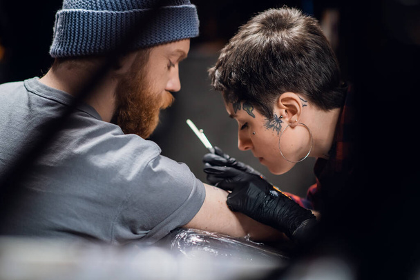 Un tatuatore con capelli corti e piercing si prepara a tatuarsi su un braccio di un uomo in un salone di tatuaggi. - Foto, immagini