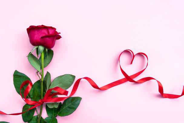 Red rose with red ribbon in shape of heart on pink background. Concept of Valentine's day, mother's day, women's day and birthday. Greetings card. Copy space. Flat lay. - Фото, изображение