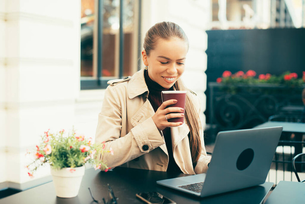 Щаслива молода жінка п'є капучино і дивиться на ноутбук, сидячи в кафе на вулиці
 - Фото, зображення