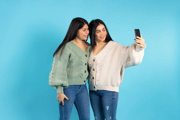 The twin sisters smile and take a selfie on a mobile phone. Blue background and empty side space. High quality photo - Foto, Bild