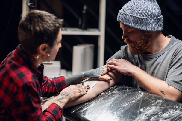 Fiatal informális lány tetováló művész tesz egy kötést vazelin a kezét egy fiatal férfi sikeres alkalmazása után egy tetoválás a kezét. - Fotó, kép